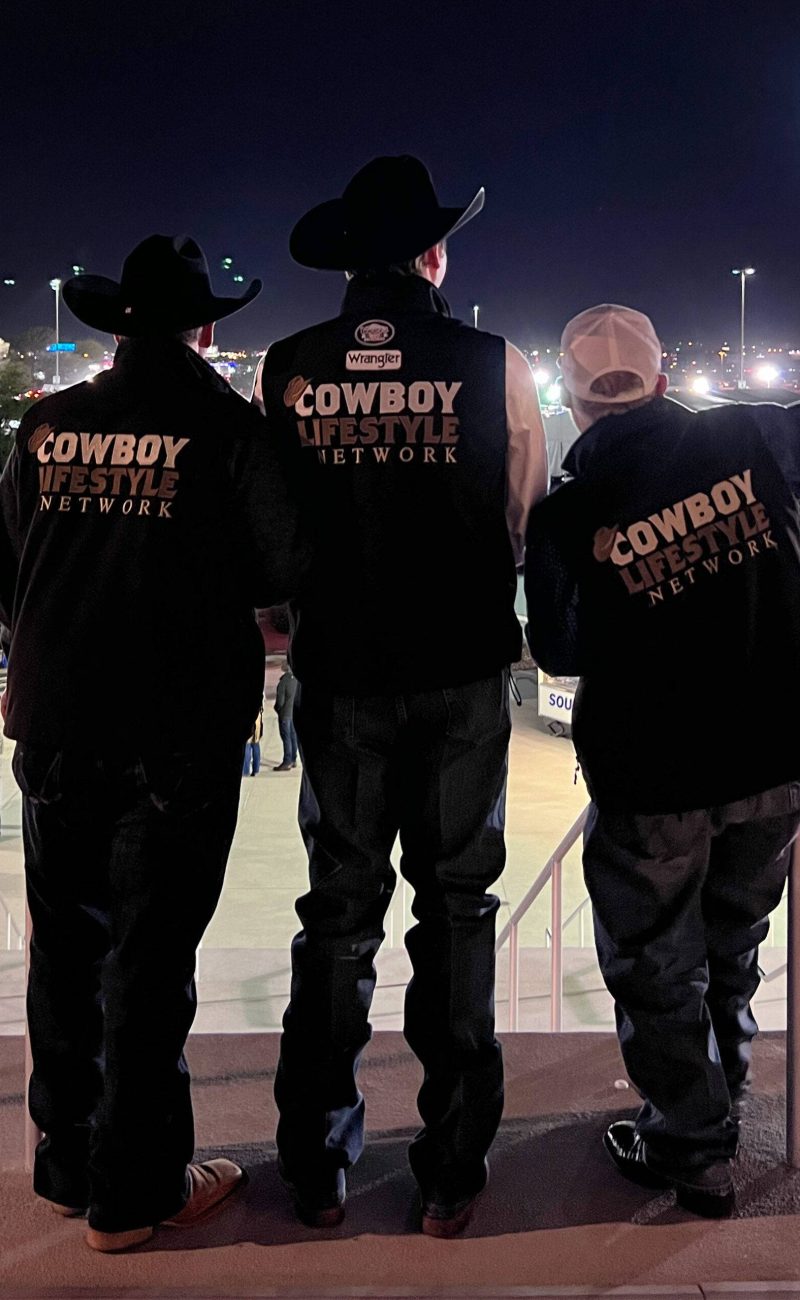 Three members of the CLN team wearing 'Cowboy Lifestyle Network' branded jackets and cowboy hats stand on a balcony overlooking a well-lit event venue at night, with a bustling crowd and bright city lights in the background.